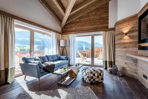 A cozy mountain lodge living room with wooden beams, plush seating, and large windows framing scenic alpine views, ideal for travelers seeking relaxation in a rustic retreat.