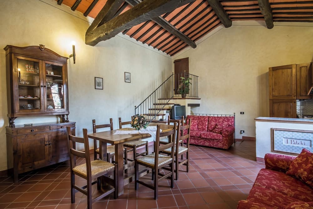 A cozy rustic living room with terracotta flooring, wooden beams, a vintage dining set, and red patterned sofas, ideal for travelers seeking a warm, traditional European retreat.