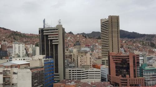 A bustling urban skyline with modern high-rise buildings and traditional red-roofed structures set against a backdrop of rugged mountains, evoking a vibrant city atmosphere ideal for cultural exploration and architectural appreciation.