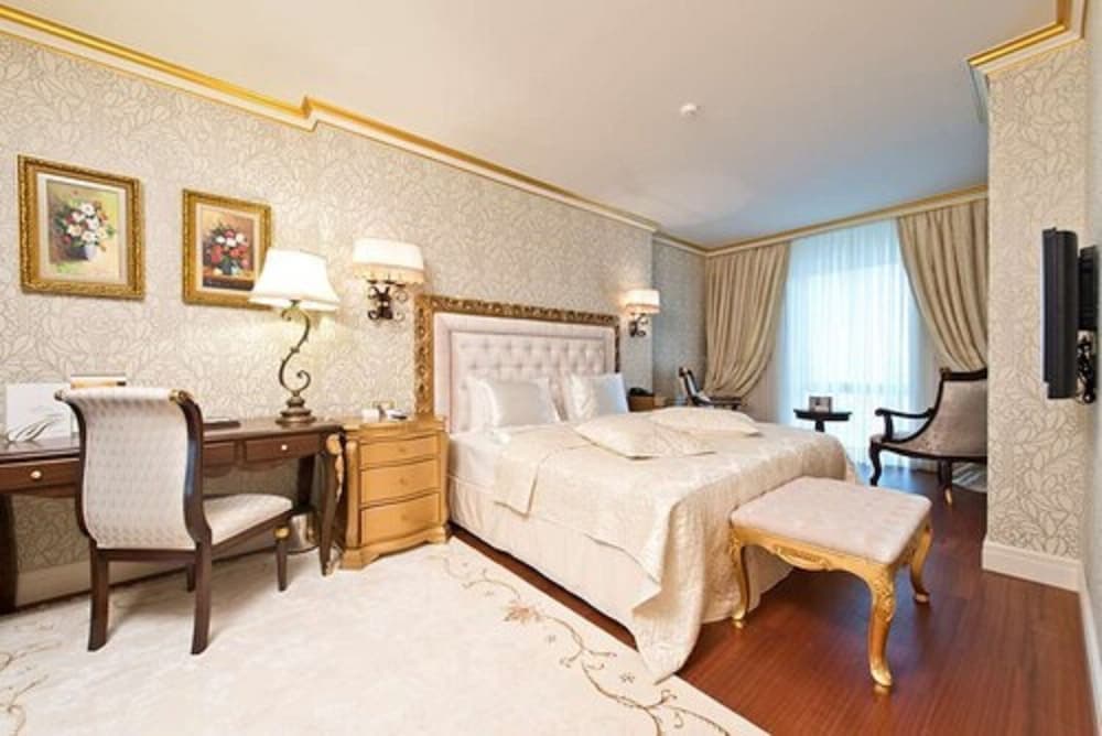 A luxurious hotel room features elegant gold-trimmed decor, plush bedding, a cozy desk area, and soft lighting, ideal for travelers seeking comfort and sophistication.