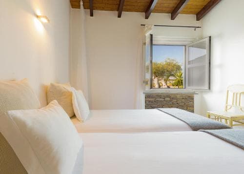 A cozy Mediterranean-style bedroom with white walls, wooden beams, and a stone windowsill offers a serene view of lush greenery, ideal for travelers seeking relaxation in a charming retreat.