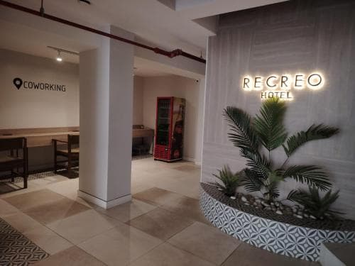 A modern hotel lobby featuring the illuminated "RECREO HOTEL" sign, a cozy coworking area, vibrant greenery in a patterned planter, and a vending machine, ideal for travelers seeking a blend of relaxation and productivity.