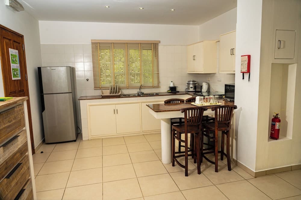 A cozy, well-lit kitchen with tiled floors, wooden blinds over the window, and a dining table set for four, ideal for travelers seeking a self-catering accommodation experience.