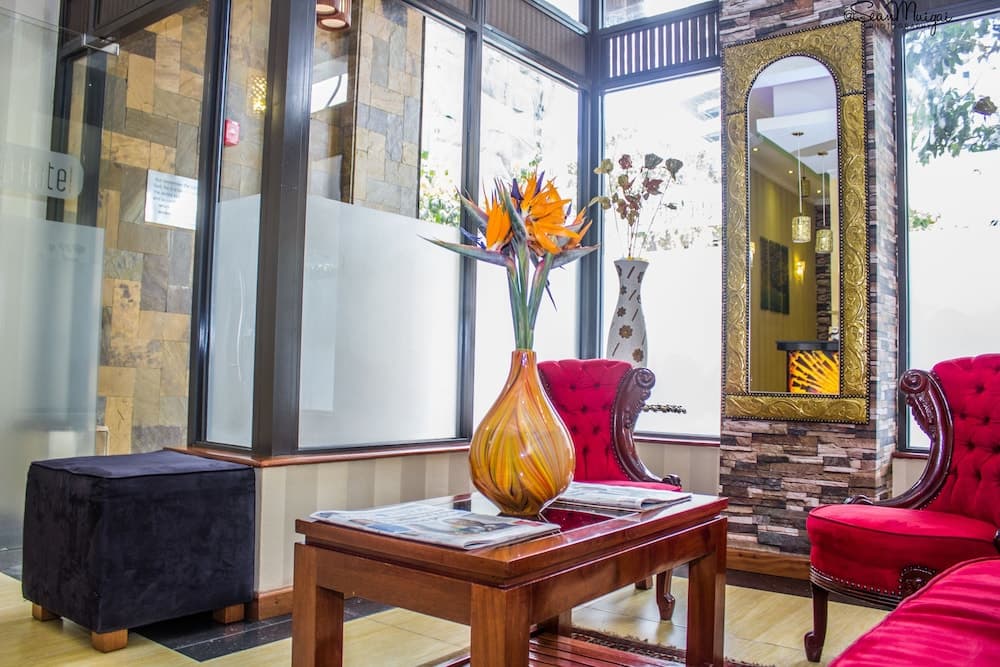 A cozy hotel lobby features plush red armchairs, a wooden coffee table adorned with vibrant flowers in an orange vase, and large windows letting in natural light, creating a welcoming atmosphere ideal for travelers seeking relaxation.