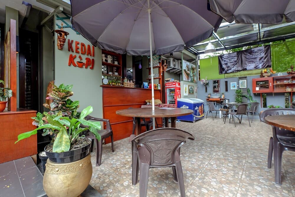 A cozy outdoor café with "KEDAI KOPI" signage offers a relaxed dining experience under large umbrellas, surrounded by potted plants and rustic decor, ideal for travelers seeking a local coffee spot.