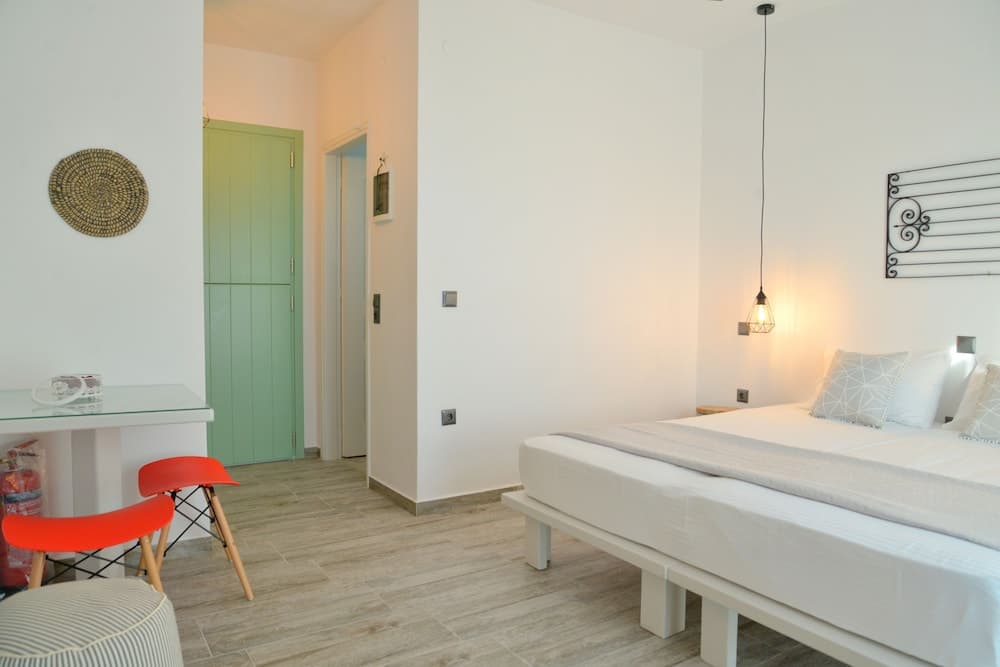 A cozy hotel room features a minimalist design with a white bed, vibrant red chairs, a green door, and warm lighting, ideal for travelers seeking comfort and simplicity.