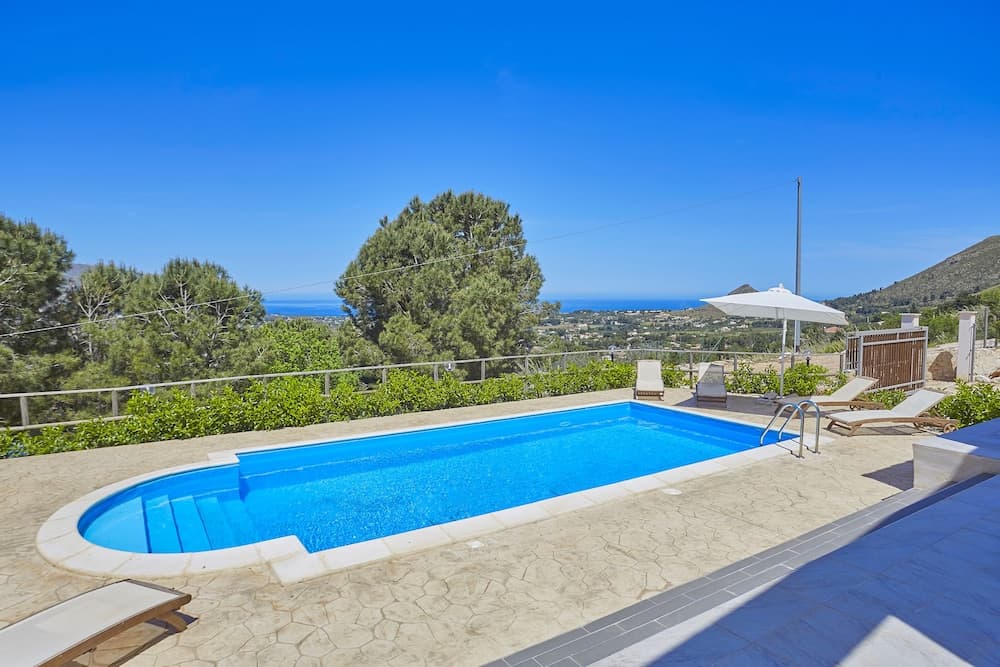 A serene private villa pool area with lounge chairs and an umbrella overlooks lush greenery and distant hills under a clear blue sky, ideal for luxury vacation retreats.