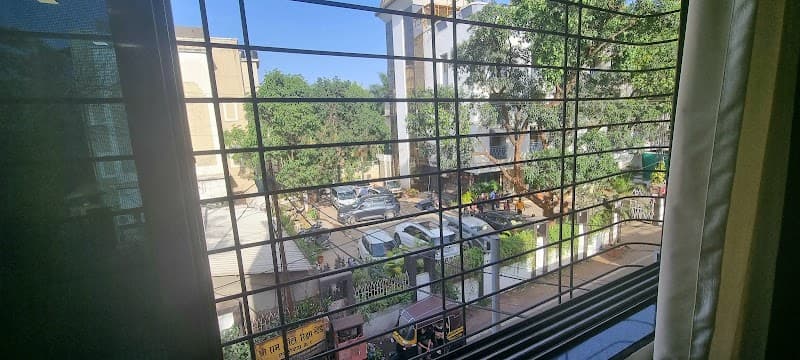 A serene view from a window with metal grills overlooks a quiet residential area with parked cars and lush greenery, ideal for travelers seeking a peaceful stay near the bus stand in Lonavala, Maharashtra.
