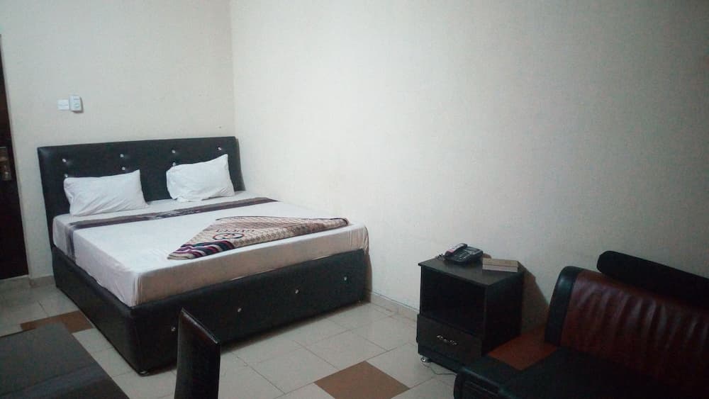 A cozy hotel room features a neatly made bed with white linens, a dark leather headboard, a small nightstand with a phone, and a leather chair, ideal for travelers seeking simplicity and comfort.