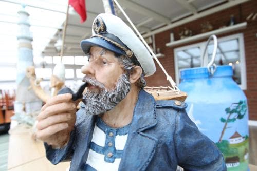 A life-sized sailor statue with a pipe and nautical attire stands in front of a brick building adorned with maritime decorations, creating a whimsical seaside atmosphere ideal for family-friendly coastal tourism.