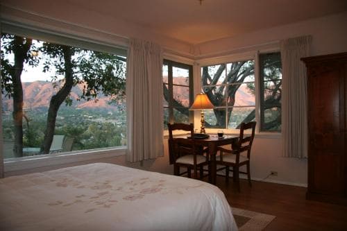 A cozy bedroom with a scenic view of rolling hills through large windows, featuring a comfortable bed, wooden furniture, and warm lighting, ideal for a relaxing getaway.