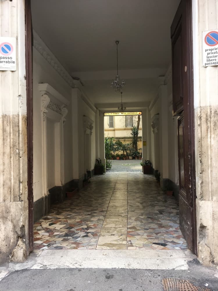 A serene passageway with mosaic-tiled floors, ornate columns, and hanging chandeliers leads to a tranquil courtyard, ideal for exploring historic European architecture and hidden urban oases.
