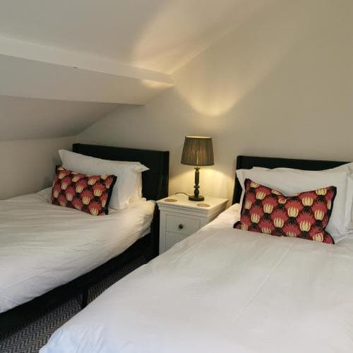 A cozy attic bedroom with two neatly made beds adorned with vibrant patterned pillows, a small bedside table with a lamp casting warm light, ideal for travelers seeking a charming and intimate accommodation experience.