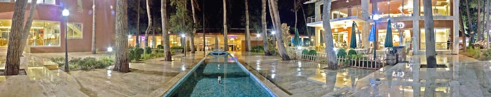 A serene evening at a luxurious resort features a glowing pool surrounded by tall trees and warmly lit buildings, ideal for travelers seeking relaxation and upscale amenities.