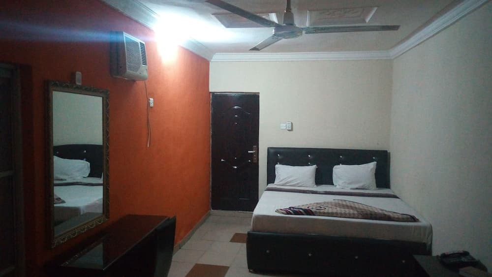A cozy hotel room features a neatly made bed, a mirror reflecting the space, an air conditioner, and warm lighting, ideal for travelers seeking comfort and simplicity.