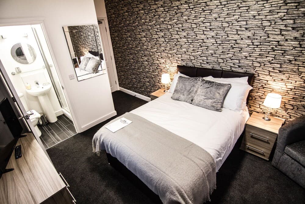 A cozy hotel room features a neatly made bed against a textured stone wall, warm bedside lamps, and an en-suite bathroom, ideal for travelers seeking comfort and style.