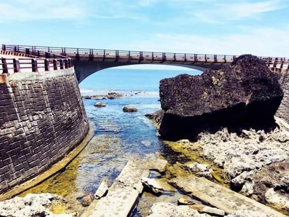 A serene coastal scene features a stone bridge arching over calm waters, surrounded by rugged rocks and a clear blue sky, ideal for tranquil seaside exploration and photography.