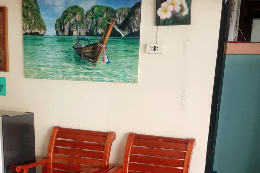 A cozy indoor waiting area features wooden chairs beneath a vibrant mural of a longtail boat on turquoise waters near limestone cliffs, evoking a serene tropical escape ideal for travelers seeking relaxation.