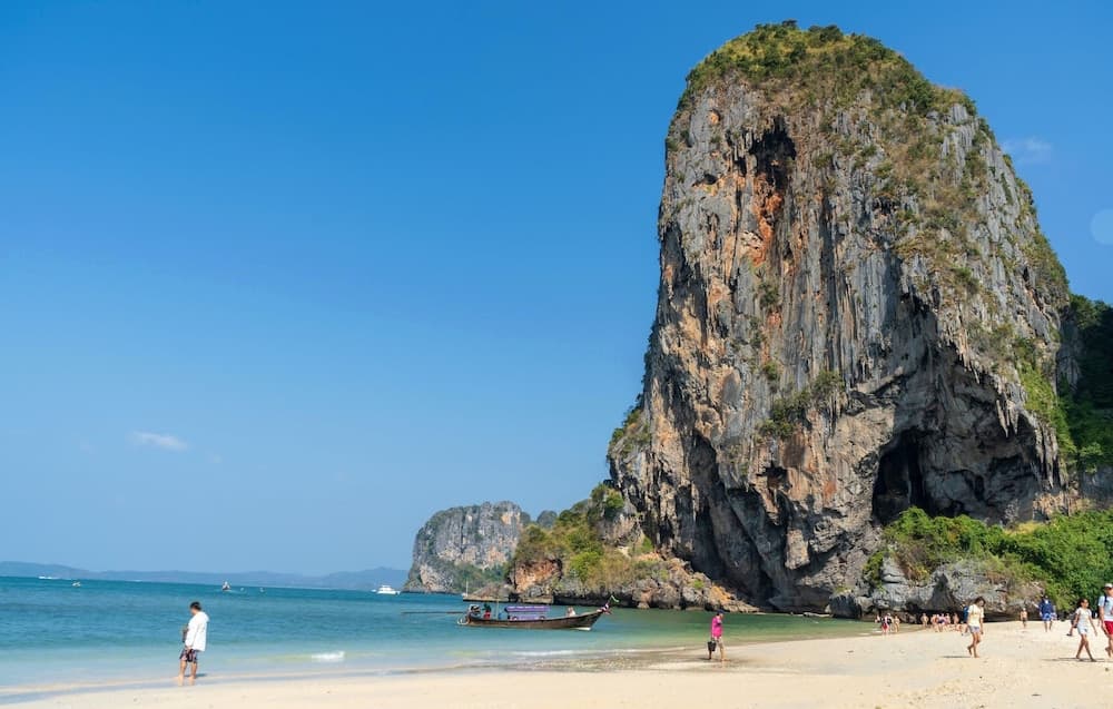 A serene tropical beach features a towering limestone cliff under a clear blue sky, with visitors enjoying the sandy shore and boats docked nearby, ideal for adventure seekers exploring coastal landscapes.