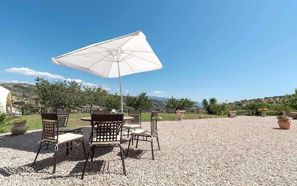 A serene outdoor patio with a gravel surface, metal chairs under a white umbrella, surrounded by lush greenery and distant hills, ideal for a relaxing vacation retreat.