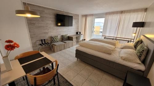 A cozy hotel suite features a modern living area with a comfortable bed, dining table, and large window offering a serene view, ideal for travelers seeking relaxation and convenience.