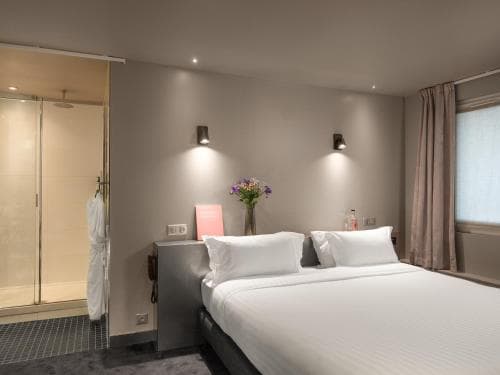 A cozy hotel room features a neatly made bed with white linens, soft lighting from wall sconces, a vase of purple flowers on the bedside table, and an open shower area, ideal for travelers seeking comfort and modern amenities.