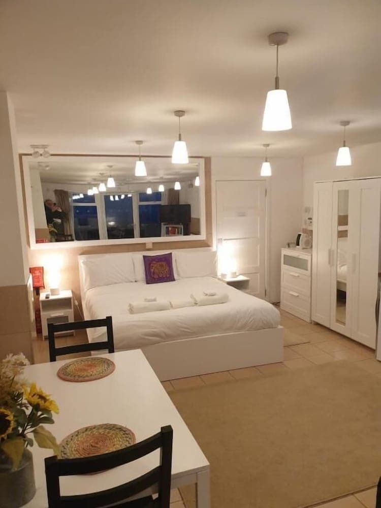 A cozy studio apartment features a neatly made bed, a dining table with sunflowers, soft lighting from pendant lamps, and a large mirror reflecting the serene ambiance, ideal for travelers seeking comfort and style in a compact living space.