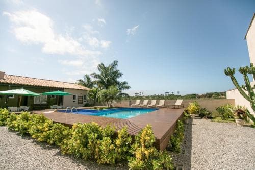 A serene tropical villa features a vibrant blue pool surrounded by lush greenery, sun loungers, and a cozy patio area under a clear sky, ideal for a relaxing vacation getaway.