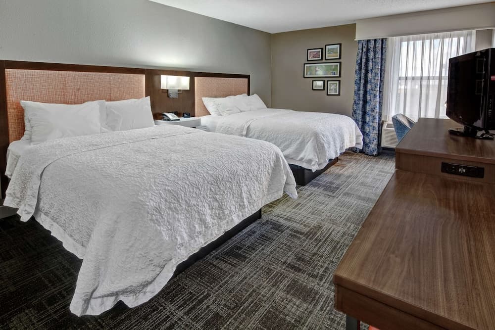 A cozy hotel room features two neatly made beds with white linens, a wooden desk, a flat-screen TV, and a window with patterned curtains, ideal for travelers seeking comfort and functionality.