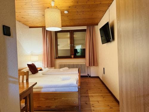 A cozy hotel room with wooden floors and ceilings, featuring a neatly made bed, a small dining table, a TV mounted on the wall, and warm lighting, ideal for travelers seeking comfort in a rustic setting.