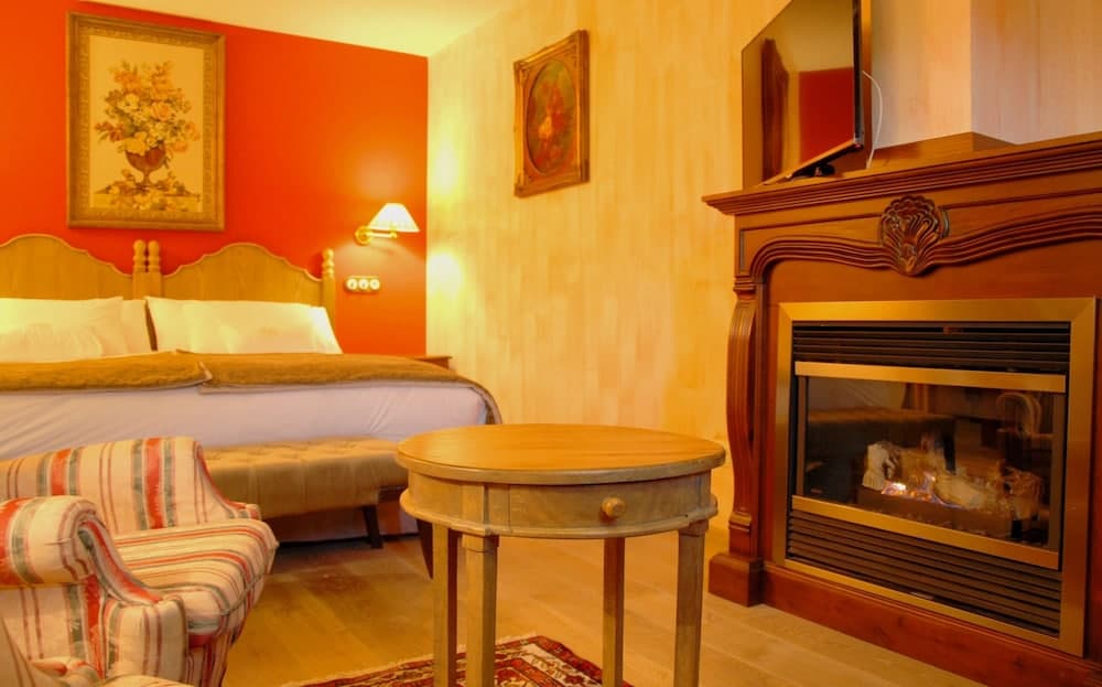 A cozy hotel room features a warm orange wall, a fireplace with a roaring fire, a wooden table, a striped armchair, and framed artwork, creating an inviting atmosphere perfect for travelers seeking comfort and relaxation.
