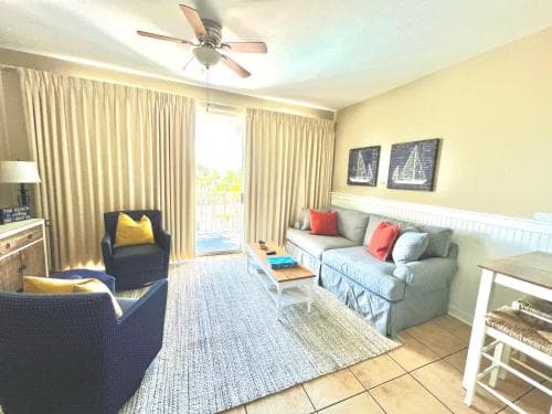 A cozy living room with a nautical theme, featuring comfortable seating, sailboat artwork, and large windows overlooking the outdoors, ideal for a relaxing beachside vacation rental.