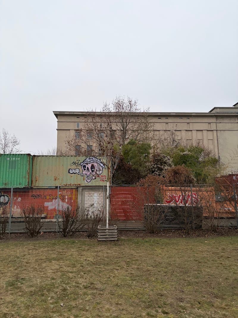 A quiet urban scene featuring graffiti-covered shipping containers and a fenced-off area in front of a large industrial building, ideal for travelers exploring Berlin's gritty, artistic side.