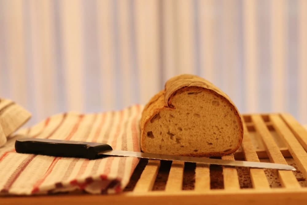 A cozy kitchen scene featuring a freshly sliced loaf of bread on a wooden cutting board with a knife, set against soft striped curtains, evokes a warm, homely mood ideal for culinary travel enthusiasts seeking authentic local dining experiences.