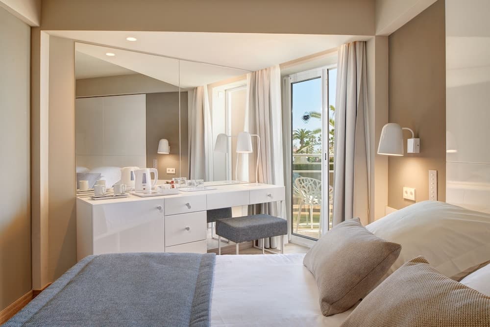 A cozy hotel room features a neatly made bed, a stylish vanity with a large mirror, and a balcony view of palm trees, ideal for travelers seeking comfort and relaxation.
