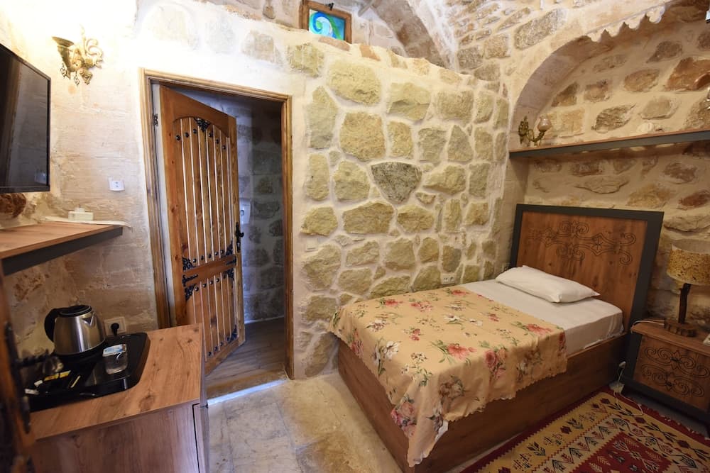 A cozy stone-walled bedroom with rustic wooden furnishings, floral bedding, and a traditional rug exudes warmth and charm, ideal for travelers seeking a unique cultural stay experience.