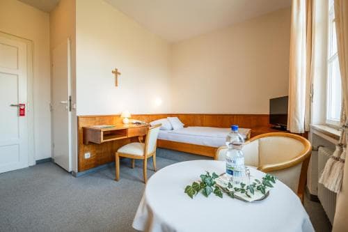A cozy hotel room with a simple bed, desk, and chair, adorned with a cross on the wall and a table set with water bottles and greenery, evokes a serene and welcoming atmosphere ideal for travelers seeking comfort and tranquility.