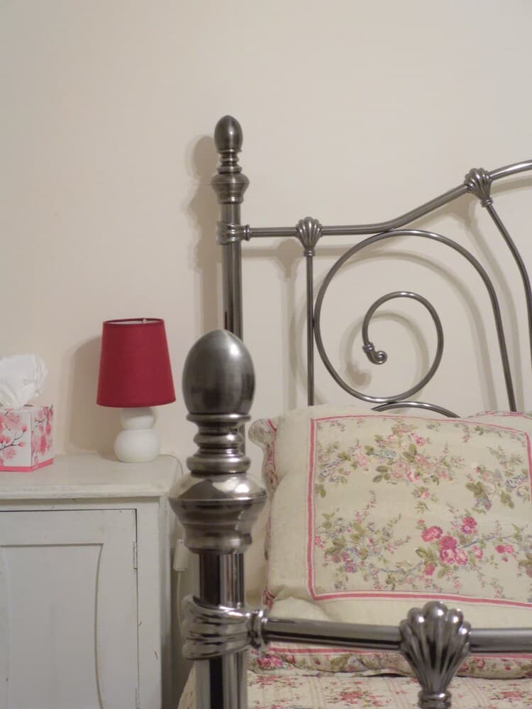 A cozy bedroom corner features an ornate metal bed frame with floral bedding, a white nightstand holding a red lampshade and tissue box, evoking a vintage charm ideal for boutique hotel stays.