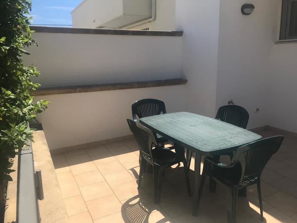 A cozy outdoor patio features a green table and chairs set against white walls, with a lush green plant adding vibrancy, ideal for travelers seeking a serene dining spot in sunny weather.