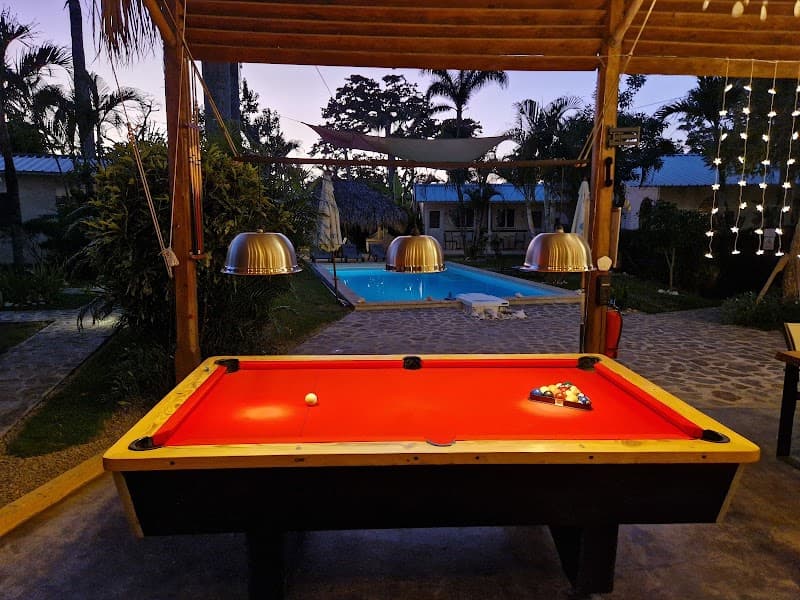 A vibrant pool table under a wooden gazebo with string lights overlooks a serene swimming pool surrounded by lush tropical greenery at dusk, creating an inviting atmosphere for leisure travelers seeking relaxation and entertainment in Las Terrenas, Dominican Republic.
