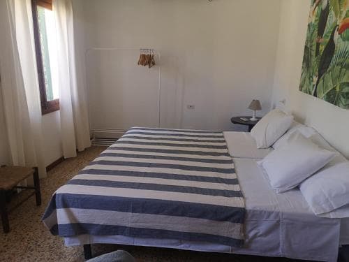A cozy hotel room features a neatly made bed with blue and white striped bedding, a small bedside table with a lamp, and a tropical-themed wall art, creating a serene atmosphere ideal for travelers seeking relaxation.