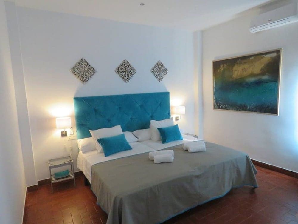 A cozy hotel bedroom features a plush bed with turquoise accents, warm lighting, decorative wall art, and terracotta flooring, ideal for travelers seeking comfort and style.