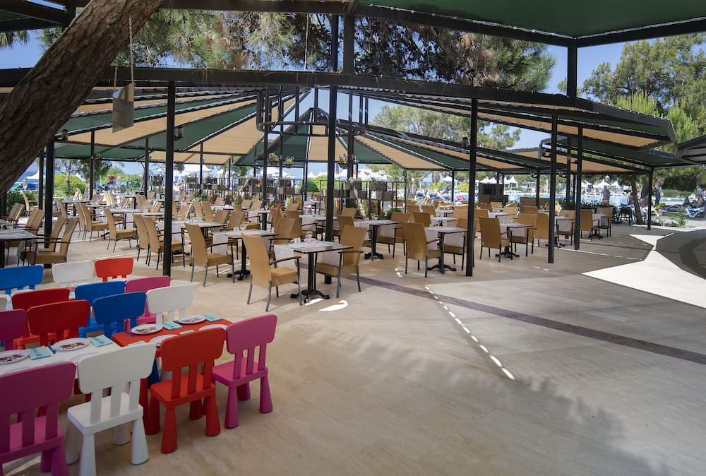 A vibrant outdoor dining area with colorful children's chairs, adult seating under green canopies, and lush trees creates a cheerful family-friendly atmosphere ideal for beachside vacations.