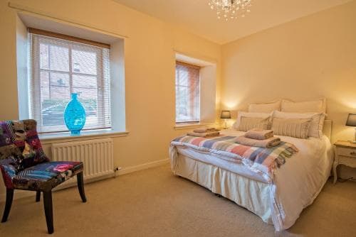 A cozy bedroom with soft lighting, a neatly made bed adorned with colorful linens, a vibrant armchair by the window, and a chandelier overhead creates an inviting atmosphere ideal for a relaxing getaway.