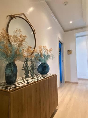 A cozy hotel hallway features a stylish console table adorned with blue vases and dried flowers under a round mirror, creating a welcoming atmosphere ideal for travelers seeking comfort and elegance.