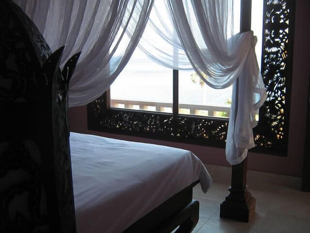 A serene hotel bedroom features a neatly made bed beside a window adorned with flowing white curtains and intricate wooden carvings, ideal for travelers seeking a peaceful retreat.