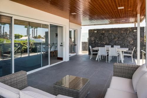 A modern patio with sleek furniture, glass doors leading to an indoor dining area, and a stone wall backdrop offers a serene and stylish retreat for travelers seeking relaxation in a contemporary villa setting.