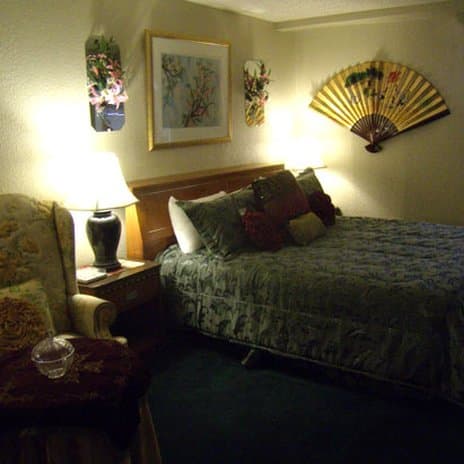 A cozy hotel room features a neatly made bed adorned with patterned bedding, warm lighting from a bedside lamp, decorative wall art including a fan, and a comfortable armchair, ideal for travelers seeking relaxation.