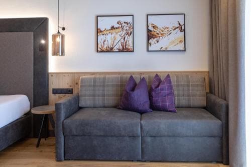 A cozy hotel room features a plush sofa adorned with purple pillows, framed mountain artwork on the wall, and warm lighting, creating an inviting atmosphere ideal for travelers seeking relaxation.