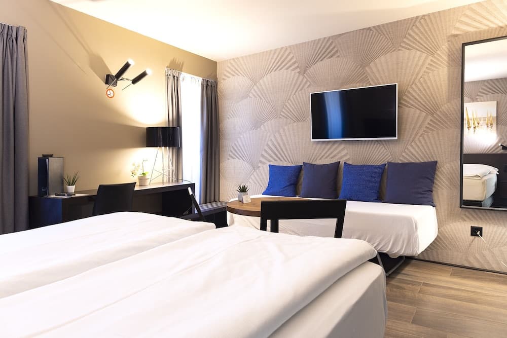A cozy hotel room features two neatly made beds, a stylish desk area with modern lighting, a wall-mounted TV, and a geometric-patterned accent wall, ideal for travelers seeking comfort and functionality.